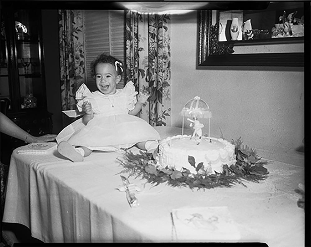 Charlene Foggie Barnett, on her first birthday, in light colored dress and hair bow, seated on table beside cake with dancer on top, in parsonage of Wesley Center Church photo by Charles "Teenie" Harris, 1959